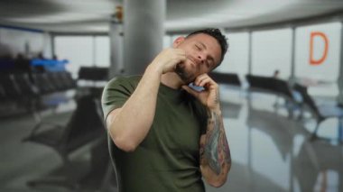 Dövmeli adam havaalanı terminalinin içinde yüz kaslarını esnetiyor. Sandalyeler ve tabelalar görünüyor..