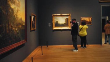 Gri duvarlarda büyük çerçeveli resimlerle çevrili loş aydınlatılmış bir müze galerisinde resim çeken bir adam..