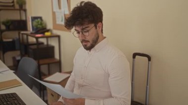 Sakallı ve gözlüklü genç adam endişeli görünüyor. Teknoloji, kitaplar ve yeşillik ile çevrili modern ofiste belgeler okuyor. Profesyonel ortamlarda stres taşıyor..