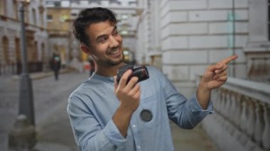 Dışarıdaki şehir merkezinde kamerayla gülümseyen ve şehir caddesini işaret eden Latin asıllı genç adam canlı sahnede fotoğrafçılık ve keşif için coşku gösteriyor.