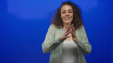 Mavi bir stüdyoda elleri sımsıkı bağlı ve gözleri kapalı bir kadın, elleri yüze yakın dua pozu, beyaz bir kolsuz tişört üzerine katıksız yeşil bir gömlek giymek; sessiz bir minnettarlık.