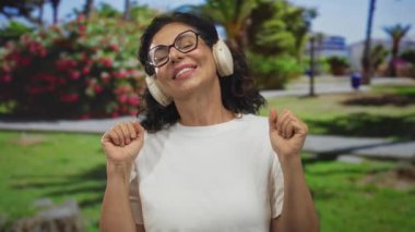 Güneşli bir parkta kulaklıkla müzik dinleyen bir kadın, yemyeşil ve çiçeklerle çevrili, gözlük takıyor ve beyaz gömlek giyiyor, gülümsüyor ve rahatlıyor..