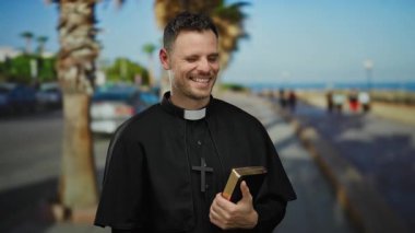 Papaz kılığında gülen bir adam, deniz kenarındaki gezinti güvertesinde incil, arka planda palmiye ağaçları ve okyanus, sıcaklık ve maneviyatı çağrıştırıyor..