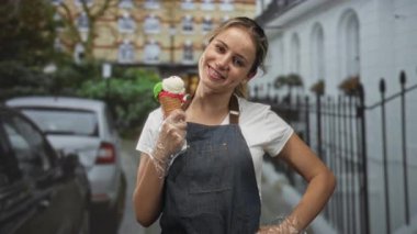 Kot önlüklü kadın, elinde eldivenli dondurma külahıyla şehir sokaklarında izleyiciye gülümsüyor..