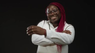 Kolları siyah bir arkaplanda çapraz gülümseyen, gözlüklü, örgülü kırmızı saçlı, tarafsız bir ortamda güven ve neşe saçan bir kadın, çeşitlilik teması için mükemmel..