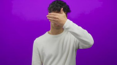 Beyaz kazak giyen genç İspanyol adam yüzünü mor stüdyo ortamında gizler; utanç.