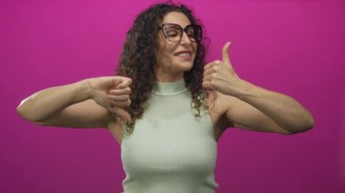 Gözleri kapalı gülümseyen genç İspanyol kadın pembe stüdyoda baş parmağını kaldırıyor ve baş parmaklarını eğiyor..
