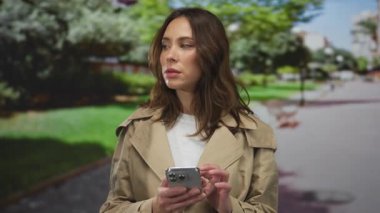 Parkta akıllı telefon kullanan, dikkatle odaklanan, yeşil ağaçlarla çevrili, modern şehir yaşam tarzı ve dijital bağlantı yayan bir kadın.