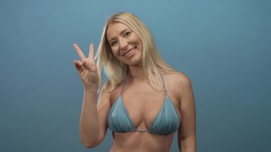 Mavi bikinili kadın gülümsüyor ve mavi stüdyoda parmakları olan barış işareti yapıyor..