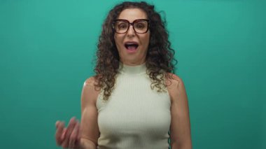 Stüdyoda gözlüklü, kolsuz yeşil bluzlu, kıvırcık saçlı, göğsüne sarılmış bir kadın..