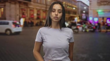 Çıplak kollu, eflatun tişörtlü bir kadın aydınlık bina cephelerinin yanındaki kalabalık bir caddede kameraya bakıyor..