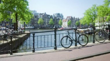 Amsterdam 'da bir kanal köprüsünde bulanık bisikletler. Açık mavi gökyüzü altında yeşil ağaçlar ve ikonik mimari içeren bir şehir manzarası var..