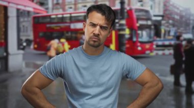 Arka planda ikonik kırmızı çift katlı otobüsle şehir caddesinde dikilen genç İspanyol adam ciddi ve düşünceli görünüyor, açık havada günlük mavi bir gömlek giyiyor..