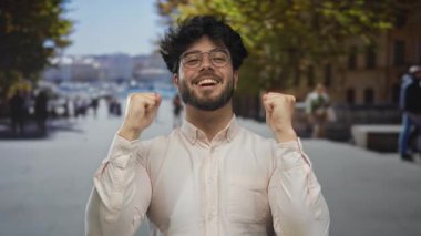 Sakallı genç adam güneşli bir sokakta kutlama yapıyor, gözlük ve beyaz gömlek giyiyor, mutluluk ve başarı yayıyor..