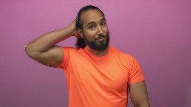 Elleri başının arkasında ve pembe stüdyoda ön kolu açık turuncu gömlekli kol kaslarını gösteren oyuncu özgüveni olan bir adam..