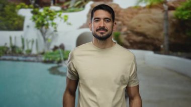 Genç İspanyol bir adam lüks bir havuz başında poz veriyor, sakin ve düşünceli bir tavır sergiliyor yemyeşil ve mimari güzelliğin ortasında..