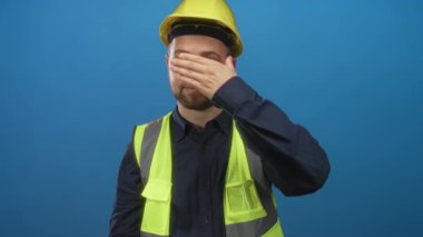 Man construction worker wearing yellow hardhat and hi vis vest covering eyes with hand in blue studio; frustration.
