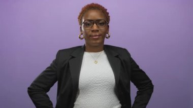 Gözlüklü ve siyah ceketli bir kadın, mor bir stüdyoda elleri kalçalarında dururken gülümsüyor; kendine güven güçlendirici güç dengesi.