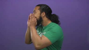 Yeşil tişörtlü bir adam mor stüdyoda elleri ağıza dayayıp yan profilde bağırıyor. Acil durum çağrısı..