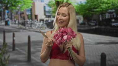 Sarışın kadın elinde pembe buket okuma kartı tutarken dekolte gösteriyor. Şehir caddesinde ışıl ışıl gülümserken. Romantik neşe..