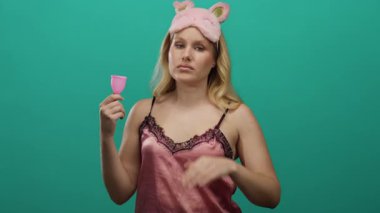 Gece elbisesi giyen bir kadın yeşil arka planda regl bardağı tutarken düşünceli görünüyor.