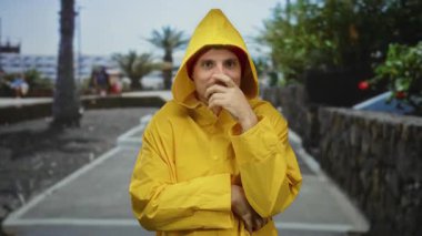 Sarı yağmurluklu adam, güneşli parkta palmiye ağaçları ve mavi gökyüzü ile kameraya gülümsüyor. Mutluluk ve açık havada rahatlama yayıyor..