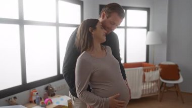 Hamile kadın ve erkek parlak bir bebek odasında kucaklaşıyorlar, umudu ve aileyi sembolize ediyorlar, şefkatli ve sevgi dolu bir ortam yaratıyorlar..