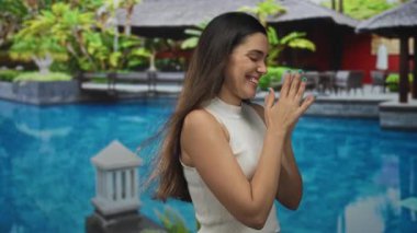 Tatil köyü havuz binasının yanında ellerini ovuşturan kadın, kolsuz üst ve deniz mavisi tırnaklar, gözleri kapalı ve öne doğru eğilmiş gülümseme; heyecan verici tatil.