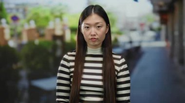 Şehir caddesinde çizgili gömlek giyen bir kadın yeşil ve bulanık arka planlı şehir ortamında düşünceli görünüyor..