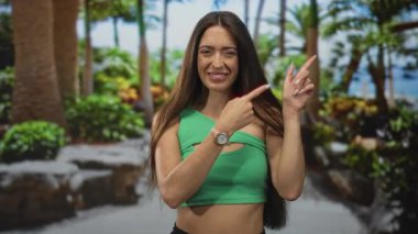 Bir ormanda dans ederken parmaklarını gösteren ve gülümseyen bir kadın. Palmiye ağaçları ve yeşil yapraklar görünür..