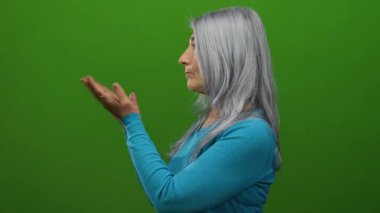 Parlak yeşil bir arka plan duvarına karşı gri saçlı yaşlı bir kadın, mavi bir gömlek giyiyor, bir stüdyo ortamında yalnızlığı ve basitliği vurguluyor..