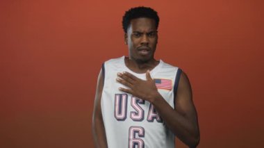Genç Afrikalı Amerikalı erkek, portakal rengi stüdyo duvarına karşı beyaz Amerikan basketbol forması giyerek göğsüne el ele tutuşuyor..