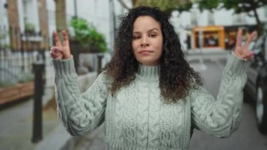 Kıvırcık saçlı, kazak giyen, şehir ortamında jest yapan bir kadın..