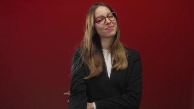 Kırmızı stüdyoda gözlüklü ve ceketli bir kadın işaret parmağıyla kamerayı işaret ediyor..