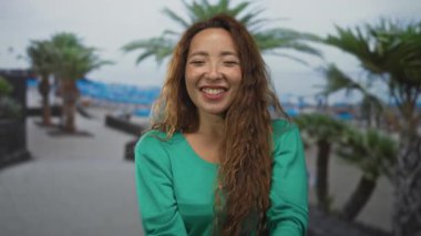 Kadın, elleri kameranın yanında olan iki parmağını ve yeşil kazakla sahil kenarında gezinen caddede gülümseyen bir gülümseme gösteriyor..