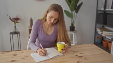 Deftere, parlak oturma odasında kahve fincanı olan bir kadın, rahat bir ortam, ahşap masa, mor gömlek, ev bitkileri, modern dekor.