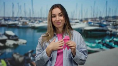 Sahilde pembe kurdeleli bir kadın kameraya gülümsüyor. Arka planda marina ve botlar var. Meme kanseri farkındalığını simgeliyor..