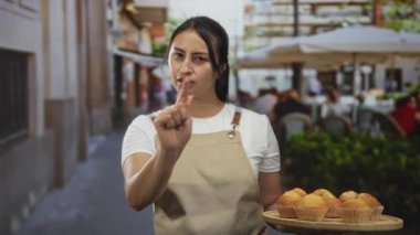 Önlüklü kadın garson, sokak kafesi terasında elinde ahşap bir tepsi kek tutarken kamerayı işaret ediyor..