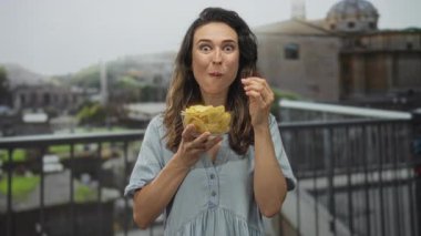 Genç İspanyol kadın güneş ışığıyla aydınlatılmış taş binanın önündeki cam kaseden cips yiyor..
