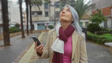 Şehir caddesinde akıllı telefon kullanan, sakin ve modern bir ortamda eşarp ve palto giyen, gri saçlı yaşlı bir kadın..