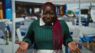 Yeşil kazaklı bir kadın açık hava restoranındaki terasta esprili bir şekilde el kol hareketi yapıyor, etkileyici vücut dili ve yüz ifadeleri sergiliyor..