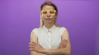 Sarı gözlüklü sarışın genç bir kadın stüdyoda mor arka plana karşı kollarını kavuşturuyor. Konsantrasyon tespiti soğukkanlılığını koruyor..