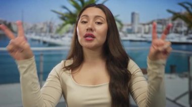Parlak güneşli deniz kıyısındaki marina binasının yakınlarındaki tırnak işareti olarak parmaklarını kullanan Latin kökenli genç bir kadın..