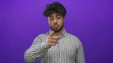 Sakallı genç İspanyol bir adam öne doğru işaret ediyor ve mor arka plan önünde çağrı yapıyor, çizgili gömlek giyiyor, iletişim ve nişan taşıyor..