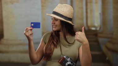 Tarihi üniversitede kredi kartıyla gülümseyen, şapka ve kamera takan, güneşli avluda el kol hareketi yapan, kültürel ortamdaki seyahat ve keşifleri kutlayan bir kadın..