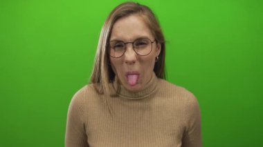 Gözlüklü kadın, izole edilmiş yeşil arka plana karşı komik yüz ifadeleri takınıyor oyuncu havasını ve günlük tarzını sergiliyor..