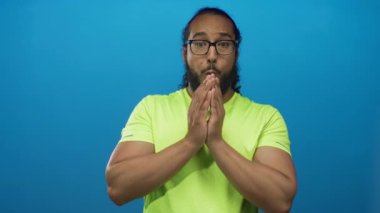 Mavi stüdyoda ağzı açık ve elleri sımsıkı kapalı, neon yeşil gömlek ve gözlük takan bir adam..