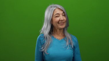 Gri saçlı yaşlı bir kadın canlı yeşil bir arkaplan karşısında neşeli bir şekilde göz kırpıyor, mavi bir gömlek giyiyor, canlı ve izole bir ortamda pozitiflik ve neşe saçıyor.