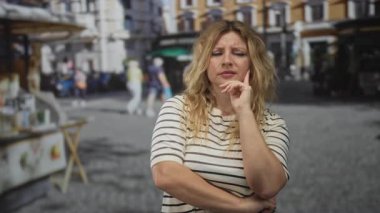 Bir sokak market tezgahı ve kaldırım taşı meydanı yanında elleri çenesinde ve kolları katlanmış bir kadın; iç gözlem kaygısı.