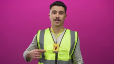 Yansıtıcı yelek giyen genç İspanyol adam, izole edilmiş pembe duvar arka planına karşı gönüllü rozeti işaret ediyor.